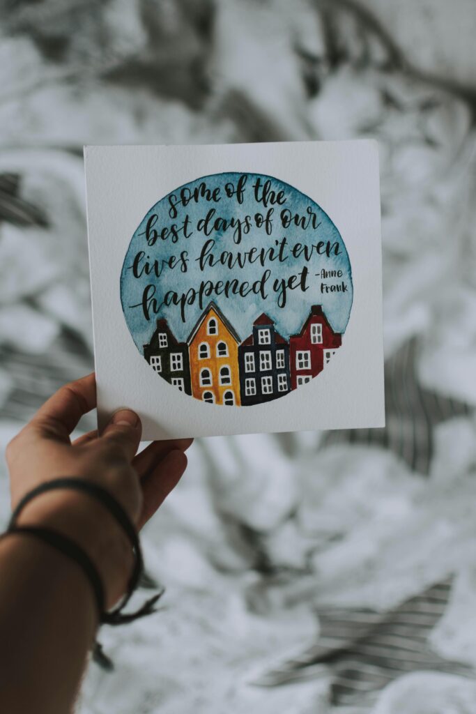 Card with an inspirational quote by Anne Frank, held in a person's hand over a blurred background.